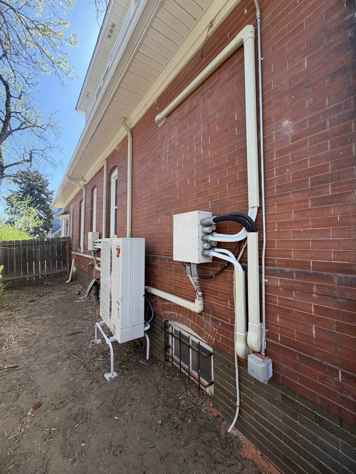 Long red brick wall with white line-set covers leading to a compact outdoor unit on a stand.