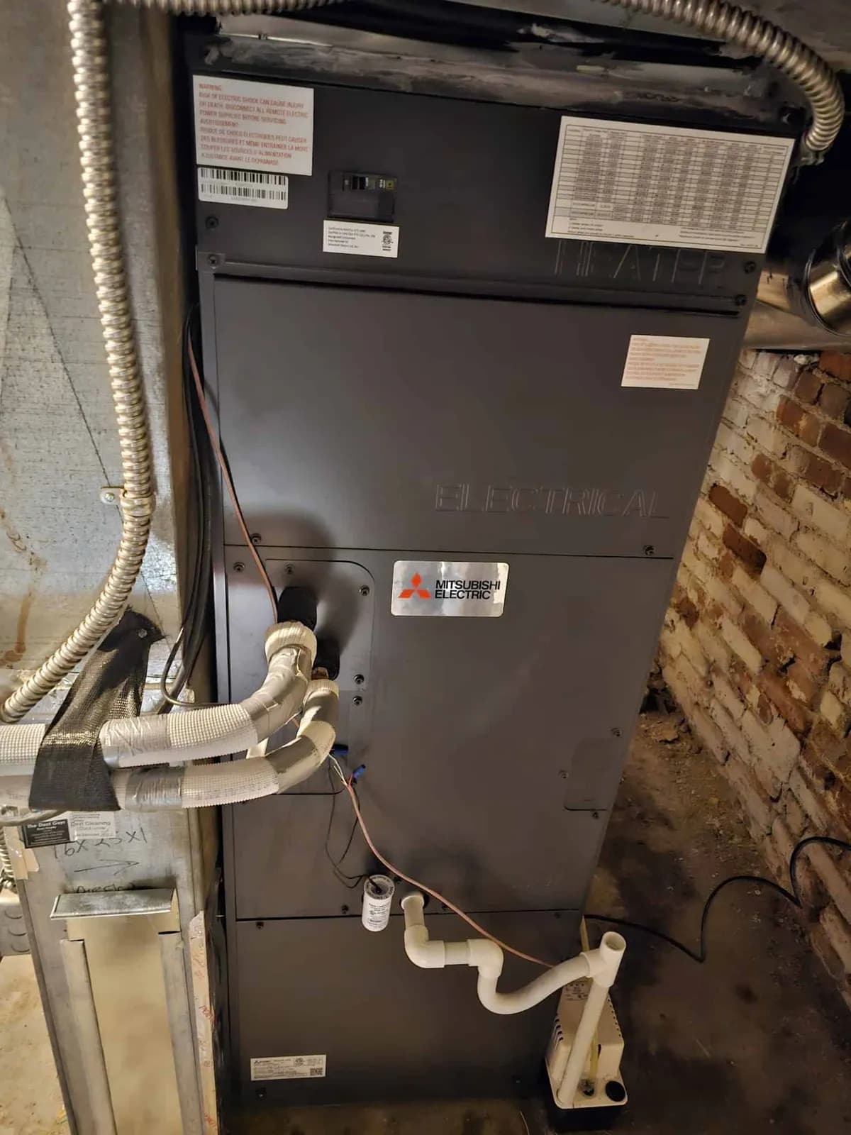 Black Mitsubishi air handler with line set by an exposed brick wall in a basement.