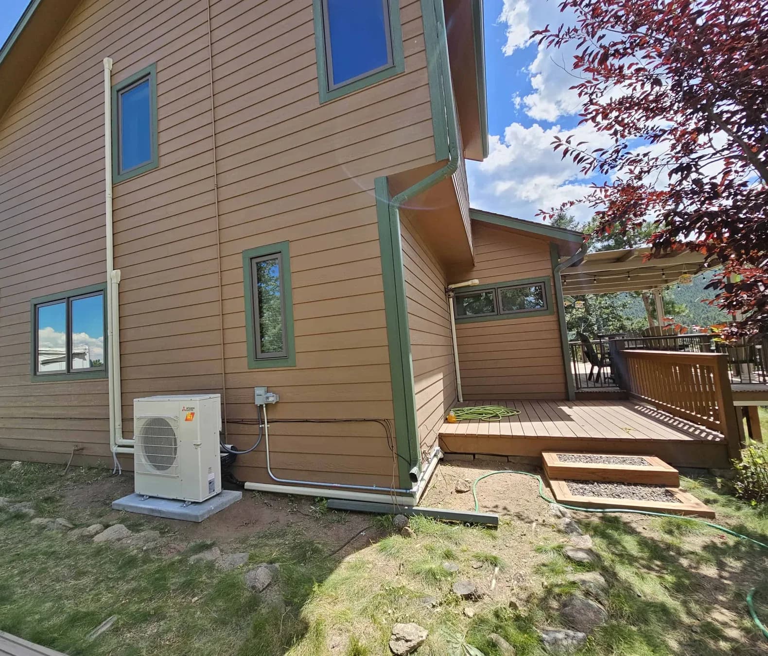 A heat pump on a pad beside a tan two-story house with a wood deck and a red-leaf tree